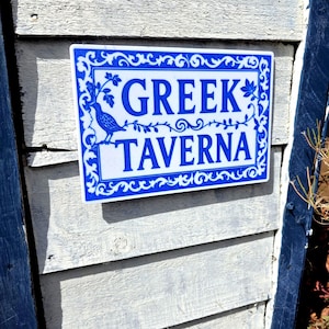 May include: A rectangular sign with a white background and blue border featuring ornate scrollwork and a bird illustration. The text GREEK TAVERNA is printed in bold blue capital letters in the centre of the sign, which is mounted on a wall.