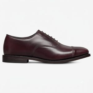 May include: A dark burgundy leather oxford shoe featuring a classic cap toe design, a low stacked heel, and a closed lacing system with thin matching laces. The shoe has a smooth, polished finish and a clean, streamlined silhouette.