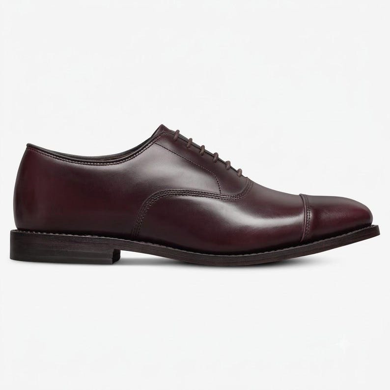 May include: A dark burgundy leather oxford shoe featuring a classic cap toe design, a low stacked heel, and a closed lacing system with thin matching laces. The shoe has a smooth, polished finish and a clean, streamlined silhouette.