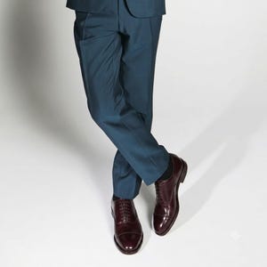 May include: A pair of dark teal trousers paired with polished dark burgundy leather oxford shoes. The trousers feature a clean, tailored cut with a straight leg, while the shoes have a classic cap-toe design with thin laces and a low heel.