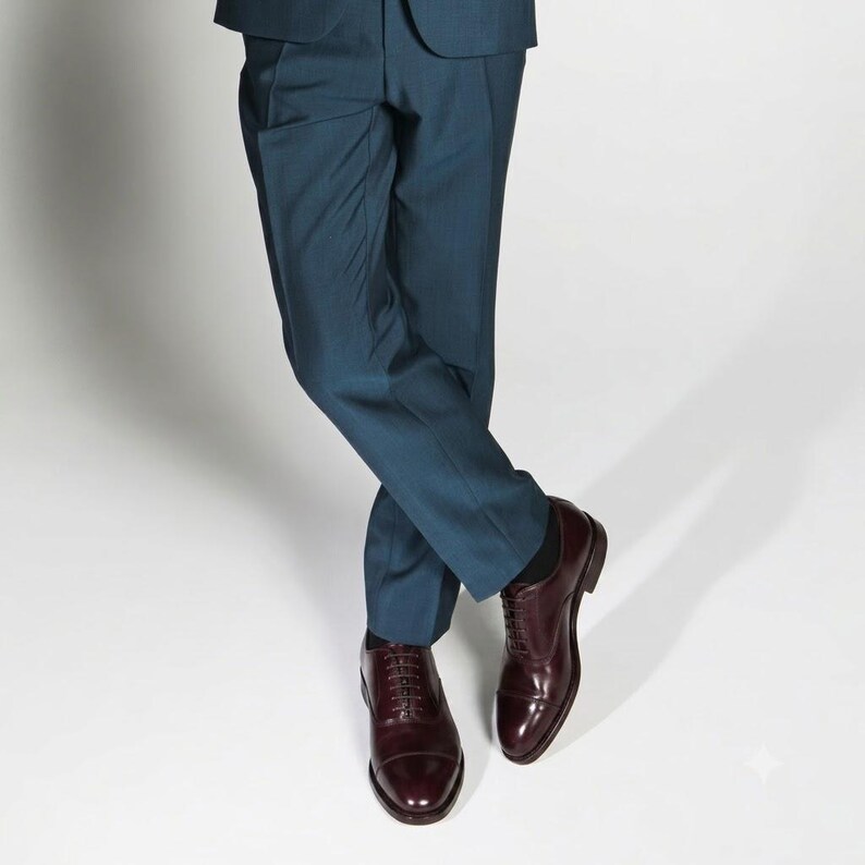 May include: A pair of dark teal trousers paired with polished dark burgundy leather oxford shoes. The trousers feature a clean, tailored cut with a straight leg, while the shoes have a classic cap-toe design with thin laces and a low heel.