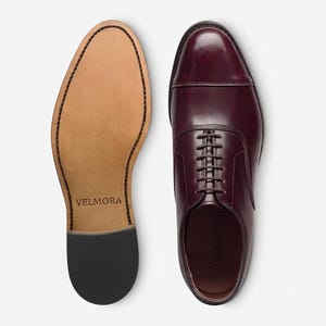 May include: A pair of burgundy leather oxford shoes with a cap toe design and dark brown laces. One shoe is shown from the top, while the other is shown from the bottom, revealing a tan sole with the brand name VELMORA embossed in the centre.