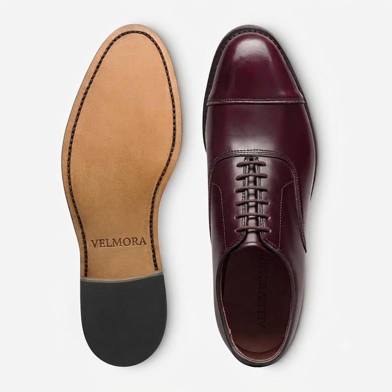 May include: A pair of burgundy leather oxford shoes with a cap toe design and dark brown laces. One shoe is shown from the top, while the other is shown from the bottom, revealing a tan sole with the brand name VELMORA embossed in the centre.