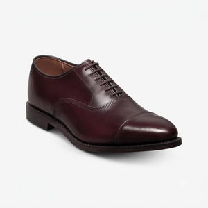 May include: A dark burgundy leather oxford shoe featuring a cap toe design, a closed lacing system with thin matching laces, a low stacked heel, and a smooth polished finish. The shoe has a rounded toe and visible stitching along the sole.