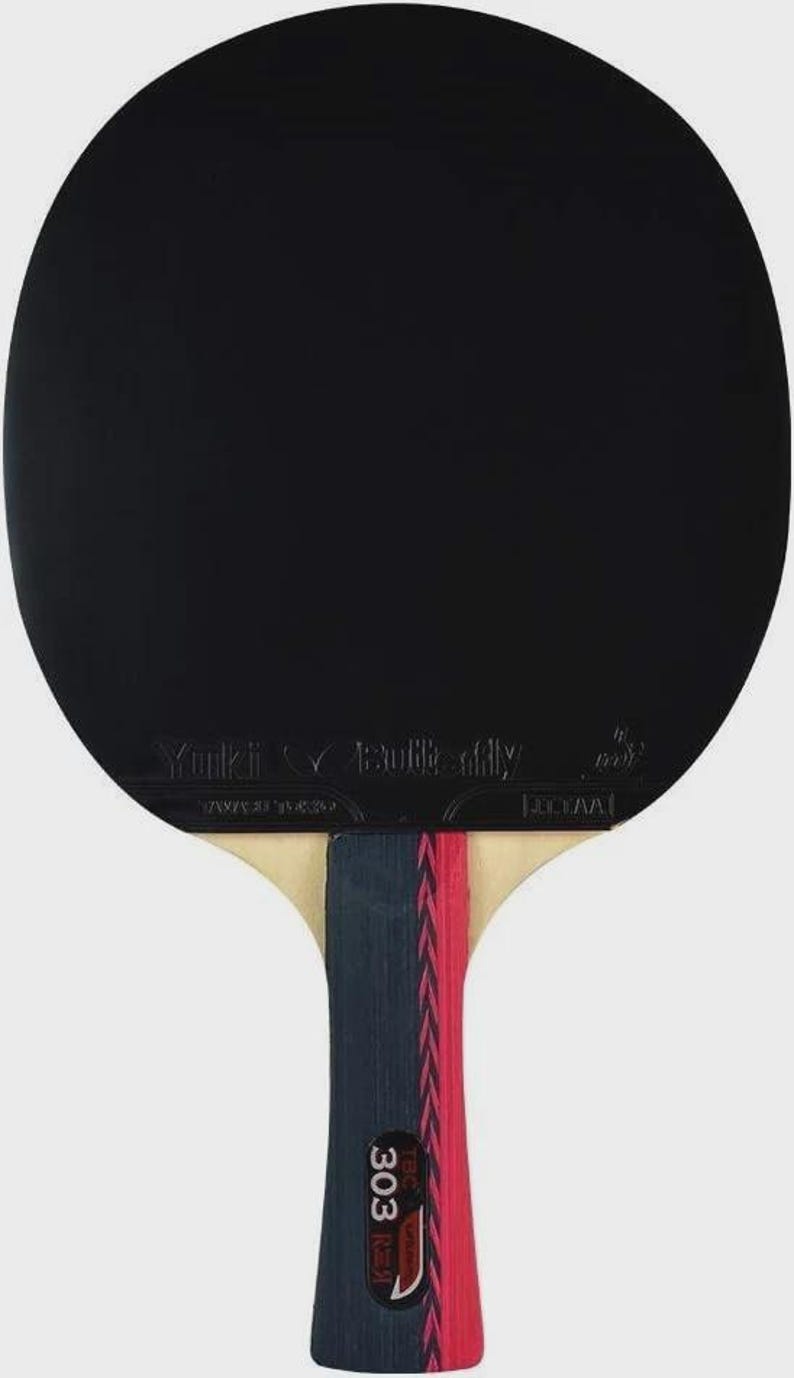 Op de afbeelding: Een zwarte tafeltennisbat met de merknaam "Butterfly" en de naam "Yuki" op het bat gedrukt. Het handvat is een combinatie van beige, marineblauw en rood. Het nummer "303" is op het handvat gedrukt.
