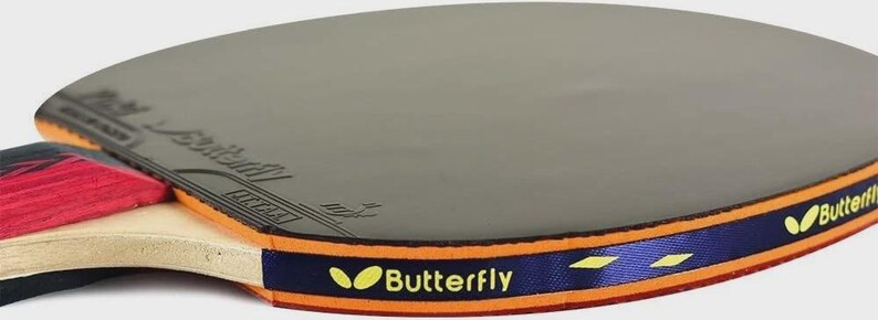 Op de afbeelding: Close-up van een Butterfly tafeltennisbat. De bat heeft een grijs speeloppervlak, een blauw en oranje rand met de merknaam en een rood en zwart handvat.