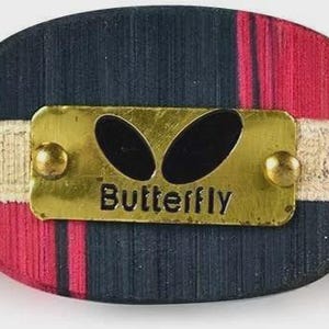 Op de afbeelding: Close-up van een tafeltennisbat-handvat. Het handvat is een mix van rood en marineblauw hout, met een goudkleurige metalen plaat met de tekst "Butterfly". Het handvat is omwikkeld met een beige band. Het blad van de bat is zichtbaar op de achtergrond.