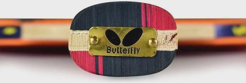 Op de afbeelding: Close-up van een tafeltennisbat-handvat. Het handvat is een mix van rood en marineblauw hout, met een goudkleurige metalen plaat met de tekst "Butterfly". Het handvat is omwikkeld met een beige band. Het blad van de bat is zichtbaar op de achtergrond.