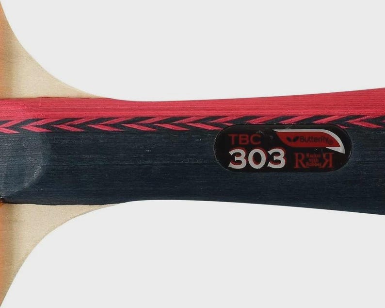 Op de afbeelding: Close-up van een zwart en rood tafeltennisbat. De bat heeft de tekst "TBC 303 R" en het Butterfly-logo. De handgreep is van hout en het ontwerp bevat rood en zwart.