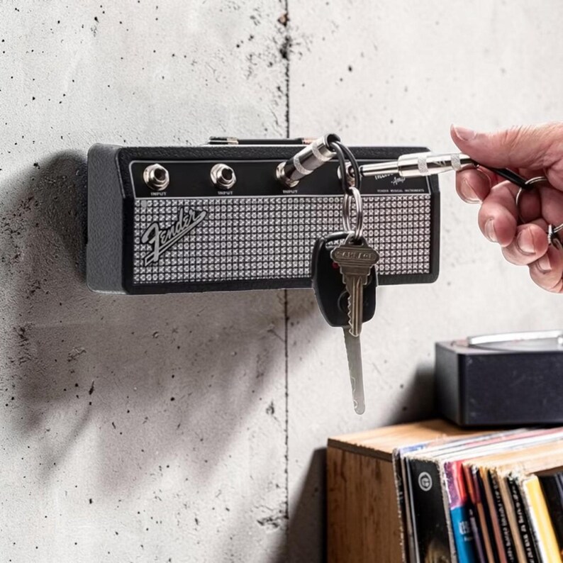 May include: A black Fender guitar amplifier wall-mounted key holder with a silver mesh front, four input jacks, and a top handle. A metal key chain plug is inserted into one of the jacks, holding a silver key and a black car key fob.