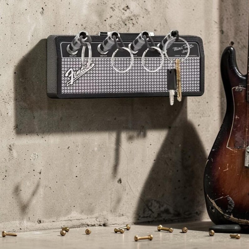 May include: A black and silver Fender Mini Twin Amp wall-mounted key holder featuring four input jacks that serve as keychains. The device has a black casing, a silver speaker grille, and the Fender logo printed on the front with key rings attached.
