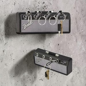 May include: A black wall-mounted key holder designed to resemble a Fender Mini Twin amplifier. It features a silver mesh front, four input jacks for keychains, and the Fender logo. The unit includes four metal keychains with plugs.