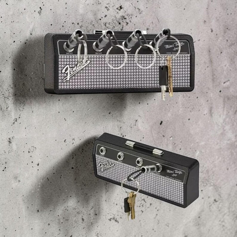 May include: A black wall-mounted key holder designed to resemble a Fender Mini Twin amplifier. It features a silver mesh front, four input jacks for keychains, and the Fender logo. The unit includes four metal keychains with plugs.