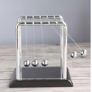 May include: A Newton's cradle desk toy with five polished steel balls suspended from a chrome frame. The base is a dark, rectangular block. The balls are suspended by thin strings, demonstrating the transfer of momentum.