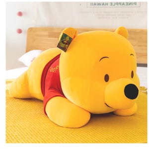 Può includere: Un grande peluche di Winnie the Pooh in posizione sdraiata. L'orso è principalmente giallo con una maglietta rossa e occhi neri. La parola "Pooh" è ricamata sulla maglietta. Un'etichetta è attaccata all'orecchio.