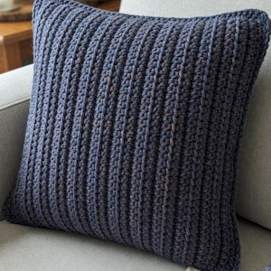 Pode incluir: Uma almofada quadrada de crochê feita à mão em tons de azul e roxo, apoiada em um sofá cinza. A almofada apresenta uma textura canelada e é feita com fio. Uma etiqueta na almofada diz "Double Crochet, Twin Pillow, Easy, 95 yds aran yarn, H/8 hook."