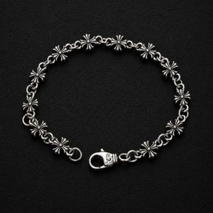 Pode incluir: Uma pulseira de corrente de metal prateado com um padrão repetitivo de pequenos elos detalhados em forma de cruz. A pulseira é fixada com um mecanismo de fecho lagosta que conecta as duas extremidades do design de corrente circular.