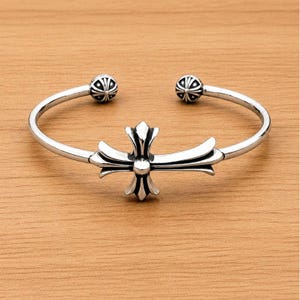 Può includere: Un bracciale a fascia in argento con un emblema centrale a croce dai bracci curvi e decorati, caratterizzato da dettagli ossidati scuri. Il bracciale aperto termina con due piccole sfere, ciascuna decorata con un motivo radiale di linee incise.