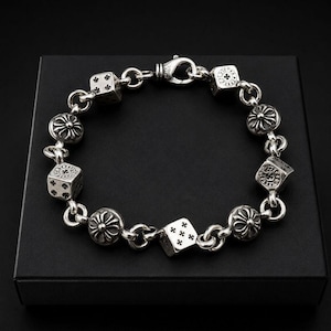 May include: A silver chain bracelet featuring alternating round charms with a cross-like floral motif and cube-shaped dice charms. The bracelet is secured with a lobster claw clasp and is constructed from polished silver links, showcasing intricate detailing.