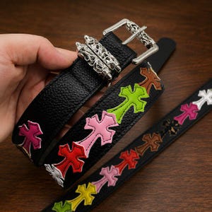 Può includere: Cintura in pelle nera con applicazioni di croci colorate. La cintura presenta una fibbia argentata con un design a teschio e dettagli decorativi argentati. Le croci sono nelle tonalità di rosa, verde, rosso e marrone.