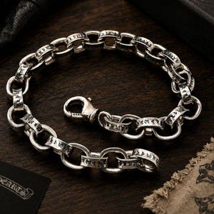 May include: A silver chain bracelet composed of thick, rounded links. Each individual link features engraved black lettering in a stylised script font. The bracelet is secured with a polished silver lobster claw clasp mechanism.