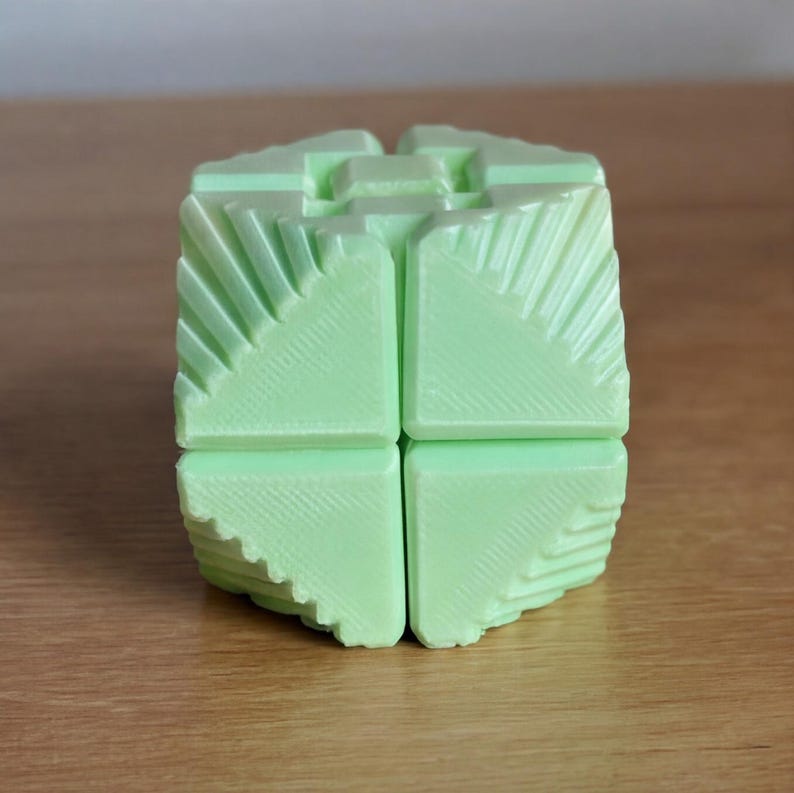 May include: A light green, geometric puzzle cube with a unique design. The cube is composed of multiple square sections, each with textured, angled patterns. The overall shape is a cube, with a complex, interlocking structure. The color is a soft, pastel green.