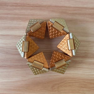 Puede incluir: Un juguete antiestrés circular compuesto por cinco segmentos triangulares interconectados. Cada segmento presenta una base metálica de color cobre con un patrón texturizado y una pieza superior triangular de color champán.