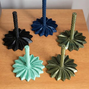 May include: A collection of five decorative candle holders in various colors. The holders feature a starburst base and a twisted, spiral-shaped stem. Colors include black, navy blue, mint green, and two shades of green. The candle holders are displayed on a wooden surface.