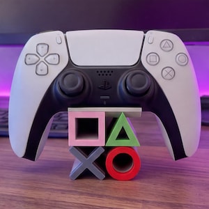 Puede incluir: Un mando de juego inalámbrico blanco y negro sobre un soporte con forma de cuatro símbolos de juego. El soporte presenta un cuadrado rosa, un triángulo verde, una cruz gris y un círculo rojo, dispuestos en una cuadrícula de dos por dos.