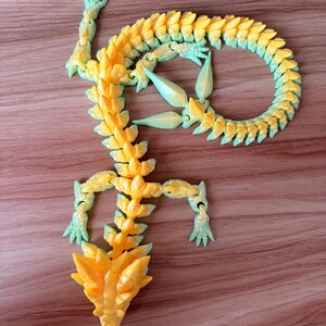 Pode incluir: Um brinquedo de dragão articulado impresso em 3D em amarelo e verde. O dragão tem um corpo, membros e cabeça segmentados, com um design detalhado e texturizado. O gradiente de cor transita do amarelo para o verde, criando uma aparência vibrante.