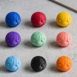 Pode incluir: Nove rolhas de garrafa esféricas e coloridas. Cada rolha é de uma cor diferente: azul, vermelho, amarelo, roxo, verde, rosa, azul claro, preto e laranja. Cada rolha tem um design de rosto estilizado e texturizado.