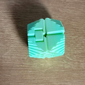 May include: A light green, geometric puzzle cube with a unique interlocking design. The cube is composed of several rectangular pieces that fit together. The surface of each piece has a textured, ridged pattern. The cube sits on a wooden surface.