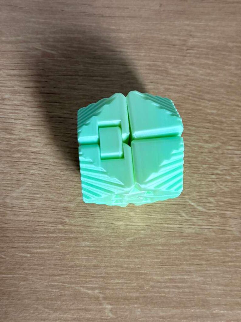 May include: A light green, geometric puzzle cube with a unique interlocking design. The cube is composed of several rectangular pieces that fit together. The surface of each piece has a textured, ridged pattern. The cube sits on a wooden surface.