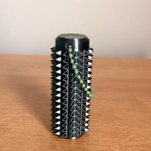 Pode incluir: Um objeto cilíndrico verde escuro com uma superfície texturizada de pontas triangulares. A parte superior e inferior são verde escuro sólido, e uma corrente de contas verdes está presa. O objeto está sobre uma superfície de madeira.