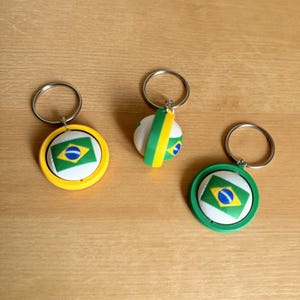 Pode incluir: Três chaveiros com a bandeira brasileira. Um chaveiro tem uma borda amarela, outro uma borda verde e o terceiro um design verde e amarelo. Cada chaveiro tem um anel prateado.