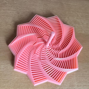 Puede incluir: Un objeto rosa, geométrico, en forma de estrella con una clavija central. El diseño presenta líneas en ángulo en capas que crean un efecto de espiral. El objeto está hecho de un material sólido y opaco y se encuentra sobre una superficie de madera.