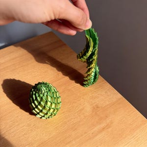 May include: A green and yellow 3D-printed geometric sculpture, with a spherical shape and a separate, elongated piece held by a hand. The objects are on a wooden surface, with shadows cast by sunlight.