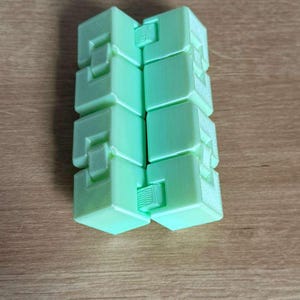 May include: A mint green infinity cube toy. The cube is made of several connected blocks with a geometric design. The toy is resting on a wooden surface.