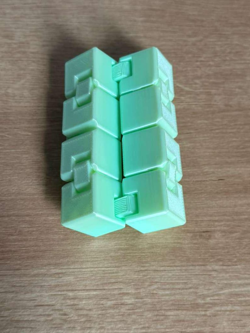 May include: A mint green infinity cube toy. The cube is made of several connected blocks with a geometric design. The toy is resting on a wooden surface.