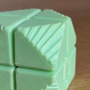 May include: A close-up of a light green cube puzzle. The puzzle is composed of several smaller cubes, each with a textured surface and diagonal lines. The overall design is geometric and modern.