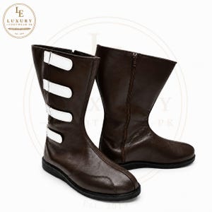 Könnte beinhalten: Ein Paar wadenhohe Stiefel in Dunkelbraun mit vier weißen horizontalen Riemen und Schnallen an der Außenseite. Die Stiefel haben eine abgerundete Zehenpartie, eine flache schwarze Sohle und einen vertikalen Reißverschluss an der Innenseite.