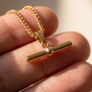 May include: A gold-toned necklace featuring a polished metal bar pendant suspended from a matching curb chain. The pendant consists of a horizontal cylindrical bar with a central spherical accent, connected to the chain by a small triangular bail.
