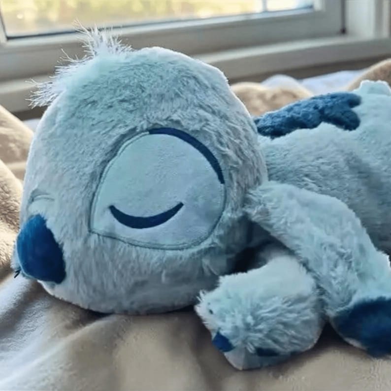 May include: A light blue plush toy of a cartoon character with large, closed eyes. The toy has a fluffy texture and is lying down, with its arms and legs extended. The character has dark blue accents on its ears, nose, and other features.