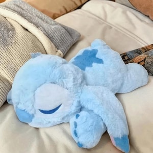 May include: A light blue plush toy in the shape of a cartoon character, lying on a bed. The toy has a smiling face with closed eyes and a darker blue patch on its back. The plush has long ears and four feet.