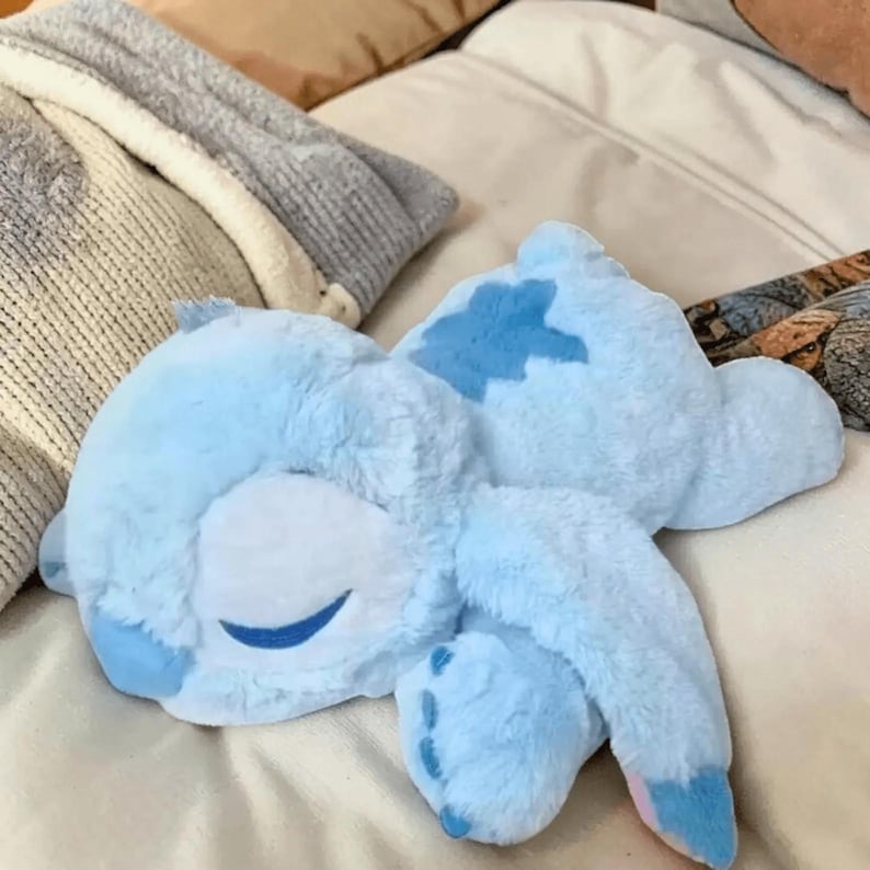 May include: A light blue plush toy in the shape of a cartoon character, lying on a bed. The toy has a smiling face with closed eyes and a darker blue patch on its back. The plush has long ears and four feet.