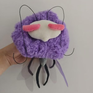 May include: A plush insect toy with a fluffy purple body, white face, and pink accents. The toy has black antennae and legs, with translucent purple wings. The toy is held in a hand.