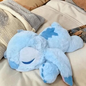 May include: A light blue plush toy in the shape of a cartoon character, lying on a bed. The toy has a smiling face with closed eyes and a darker blue patch on its back. The plush has long ears and four feet.