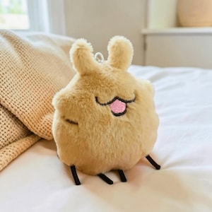 May include: A tan-coloured plush toy shaped like a creature, with a smiling face, pink tongue, and black legs. The soft toy has a fluffy texture and a small metal loop. The toy is on a white surface.