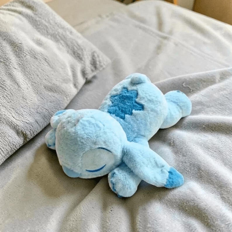 May include: A light blue plush toy in the shape of a cartoon character, lying on a gray blanket. The toy has a darker blue patch on its back and closed eyes, suggesting it is sleeping. The plush is soft and fluffy.