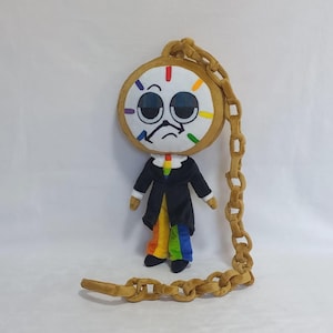 Puede incluir: Un muñeco de peluche con forma de reloj de bolsillo con una cara triste, vestido con un traje negro y pantalones arcoíris. La esfera del reloj tiene marcas coloridas y una cadena dorada. Sobre un fondo blanco.